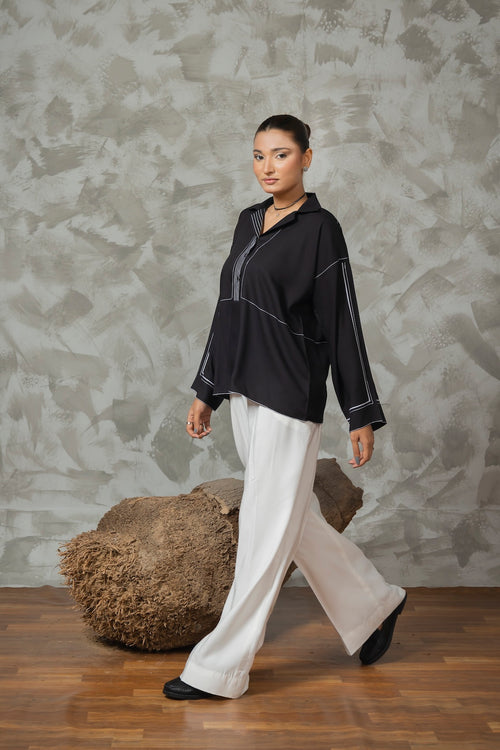 Woman wearing a black top and white pants against a textured wall.